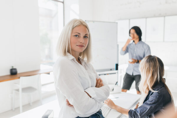 Confident blonde girl in blouse standing with arms crossed in office with big flipchart. Indoor portrait of serious female manager and her colleagues drinking coffee on background..
